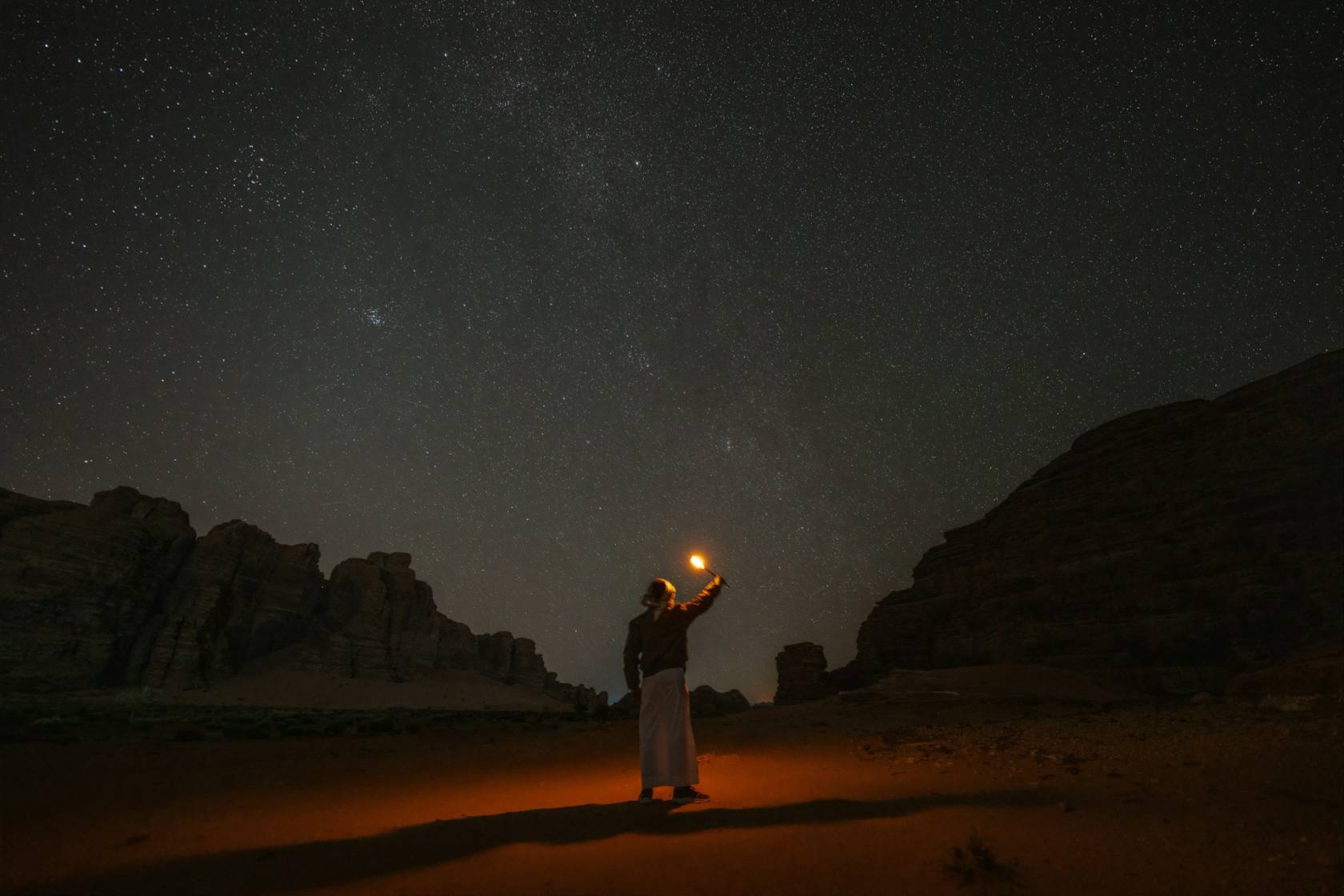 Person standing in a night landscape looking at stars, wearing clothing, under astronomical object. 